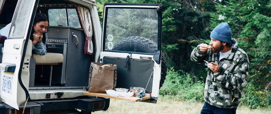 A truck with the back door open, a man eating with a beanie on